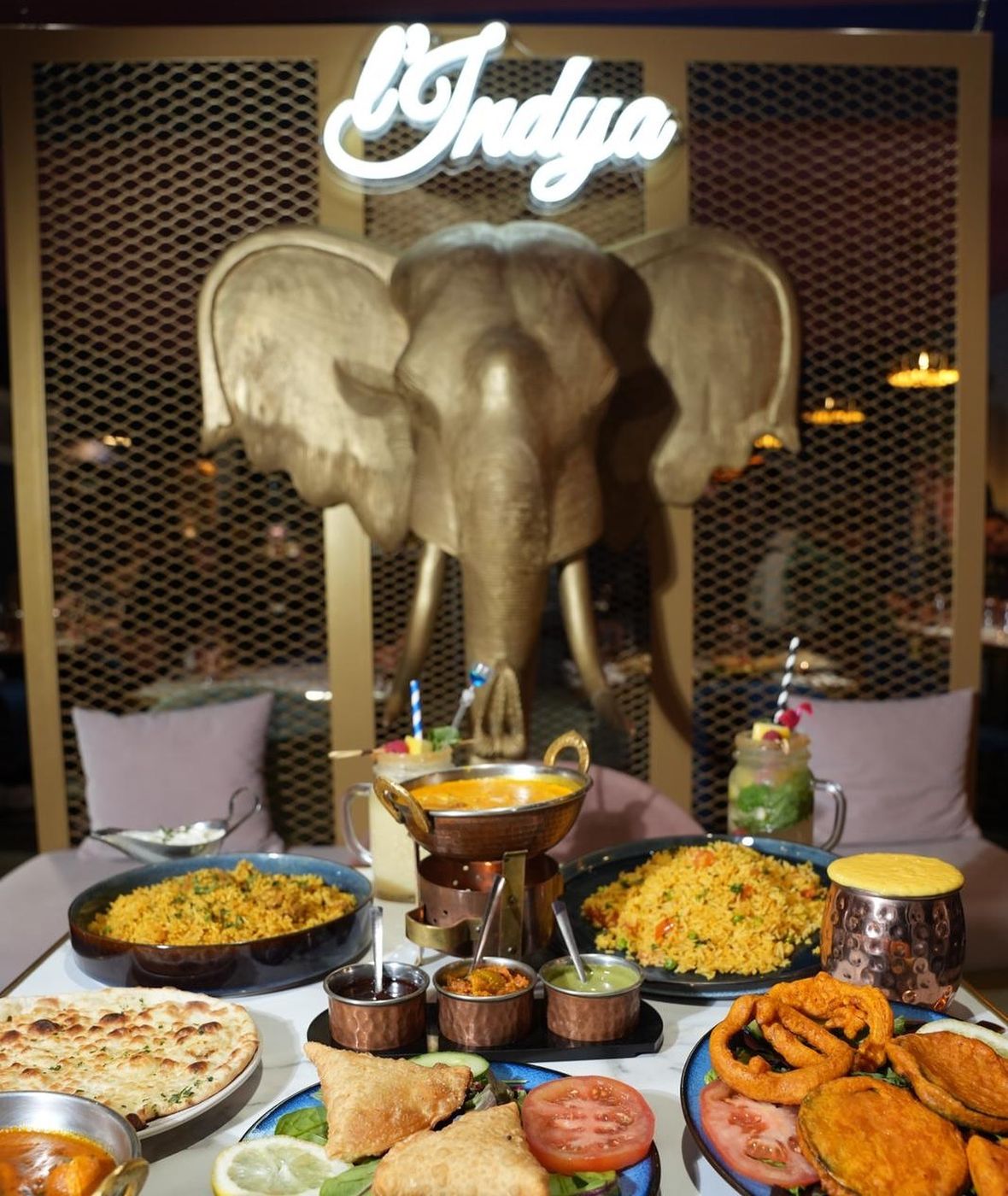 Table garnie de currys, biryanis, naans et samoussas devant l'éléphant doré et le néon L'Indya